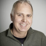 Glad man i grön tröja med kort grått hår mot grå bakgrund.