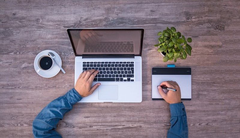 Användare vid laptop och ritplatta på träbord med kaffekopp och växt.